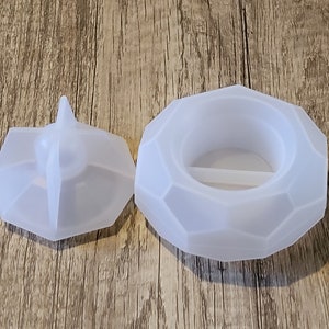 May include: A white, geometric-shaped silicone ring dish with a matching lid. The dish has a raised centre section and a small ridge around the edge. The lid has a pointed top.