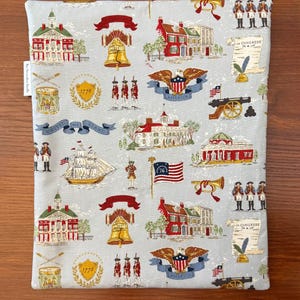 Op de afbeelding: Een lichtblauwe stoffen boekenhoes met een herhaald patroon van historische Amerikaanse afbeeldingen. Het ontwerp bevat illustraties van de Liberty Bell, gebouwen, vlaggen en soldaten. De hoes is ontworpen om boeken te beschermen.