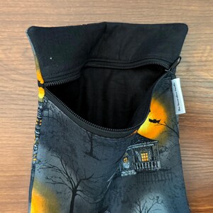 May include: A black fabric pouch with a zipper closure featuring a printed scene of a house at night with a large yellow moon, bare trees, and a bat. The interior is lined with black fabric and a white tag reads The Decorative Needle.
