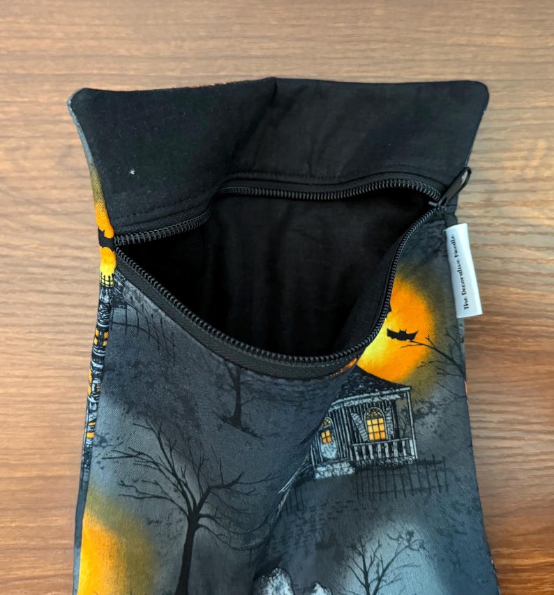 May include: A black fabric pouch with a zipper closure featuring a printed scene of a house at night with a large yellow moon, bare trees, and a bat. The interior is lined with black fabric and a white tag reads The Decorative Needle.