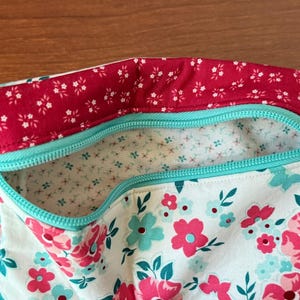 Puede incluir: Una bolsa con estampado floral con la parte superior roja y cremallera turquesa. La bolsa presenta un fondo blanco con flores rosas y turquesas. El forro interior es blanco con peque&ntilde;os estampados florales. Una etiqueta lateral dice "The Decorative Needle".