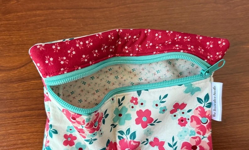 Puede incluir: Una bolsa con estampado floral con la parte superior roja y cremallera turquesa. La bolsa presenta un fondo blanco con flores rosas y turquesas. El forro interior es blanco con peque&ntilde;os estampados florales. Una etiqueta lateral dice "The Decorative Needle".