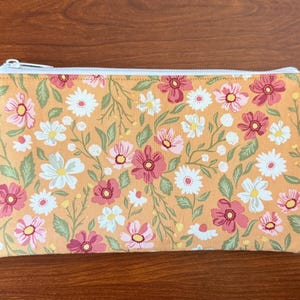 May include: A rectangular floral print pouch with a white zipper. The pouch features a pattern of pink, white, and yellow flowers with green leaves on a yellow background. The tag on the side reads "The Decorative Perdele".