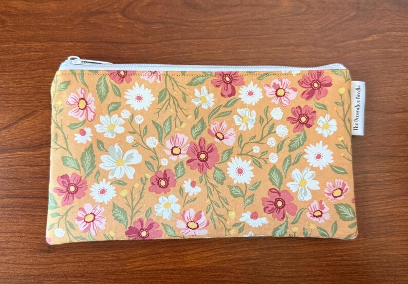 May include: A rectangular floral print pouch with a white zipper. The pouch features a pattern of pink, white, and yellow flowers with green leaves on a yellow background. The tag on the side reads "The Decorative Perdele".