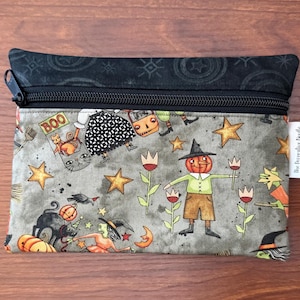 May include: A rectangular zippered pouch with a Halloween-themed pattern. The design features pumpkins, witches, cats, stars, and a scarecrow with a pumpkin head. The top and zipper are black, and the pouch has a white tag.
