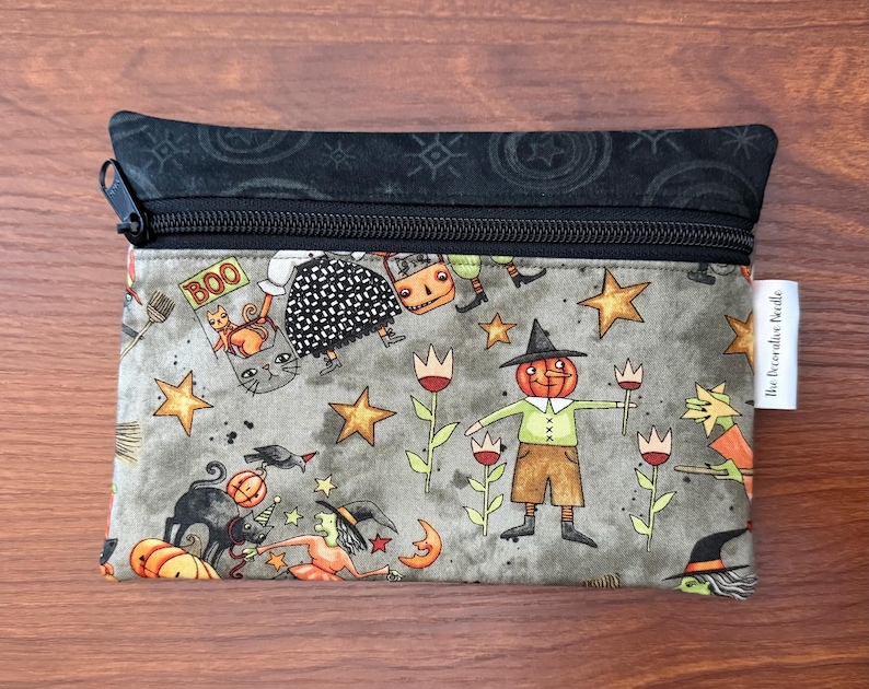 May include: A rectangular zippered pouch with a Halloween-themed pattern. The design features pumpkins, witches, cats, stars, and a scarecrow with a pumpkin head. The top and zipper are black, and the pouch has a white tag.
