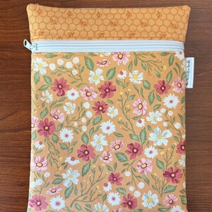May include: A rectangular zippered pouch with a floral pattern in shades of pink, white, and green on a peach background. The top section features a honeycomb pattern with bees. The zipper is white.
