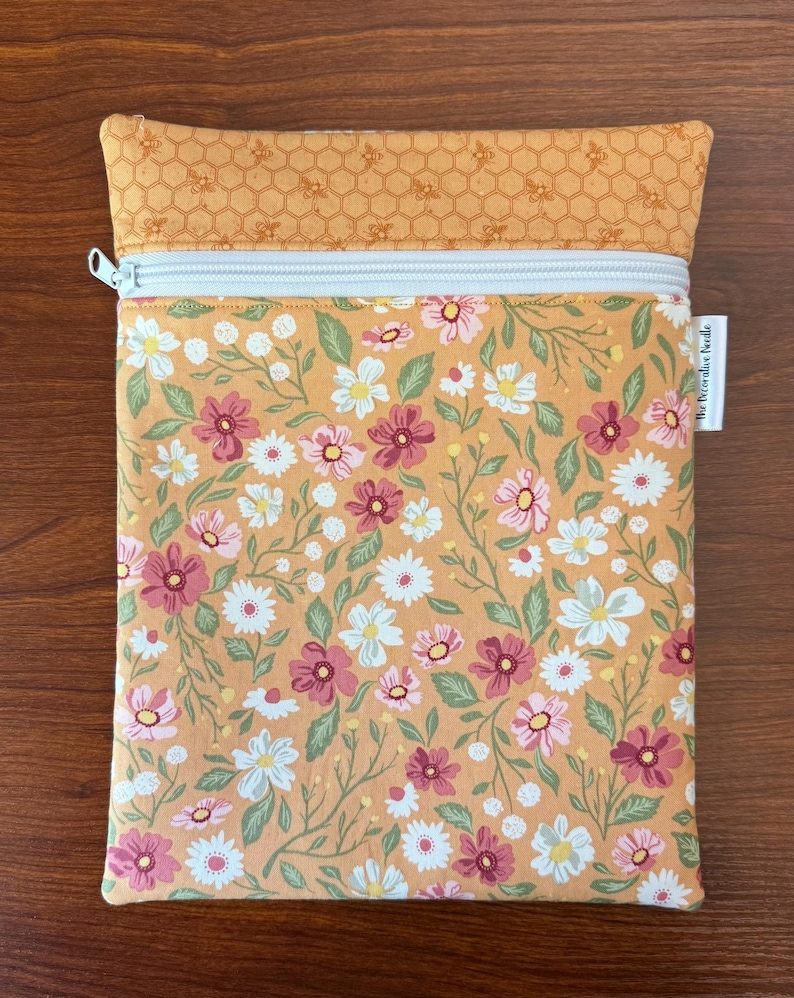 May include: A rectangular zippered pouch with a floral pattern in shades of pink, white, and green on a peach background. The top section features a honeycomb pattern with bees. The zipper is white.