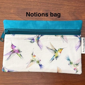 May include: A rectangular notions bag with a teal top and zipper. The bag features a white background with a pattern of colorful hummingbirds. The text "Notions bag" is printed on the top of the bag.