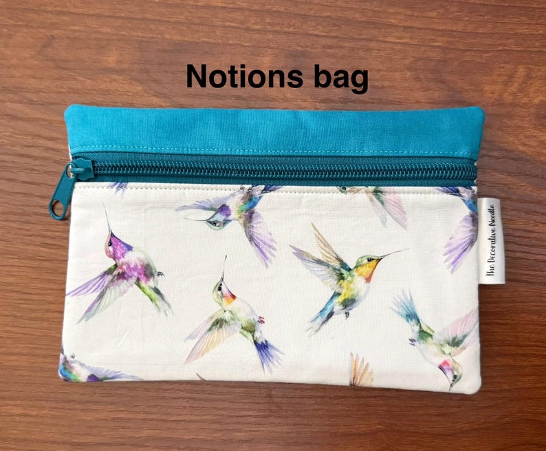 May include: A rectangular notions bag with a teal top and zipper. The bag features a white background with a pattern of colorful hummingbirds. The text "Notions bag" is printed on the top of the bag.