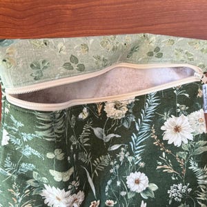 Pu&ograve; includere: Una borsa verde floreale con cerniera e una parte superiore con motivo floreale verde chiaro. La borsa presenta fiori bianchi, farfalle e fogliame. La cerniera beige &egrave; aperta, rivelando un interno bianco.
