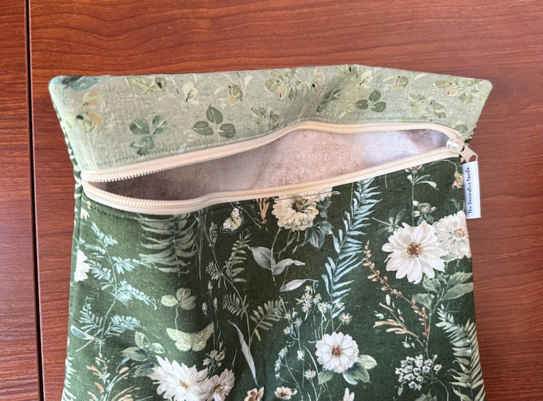 Pu&ograve; includere: Una borsa verde floreale con cerniera e una parte superiore con motivo floreale verde chiaro. La borsa presenta fiori bianchi, farfalle e fogliame. La cerniera beige &egrave; aperta, rivelando un interno bianco.