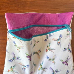 May include: A white fabric bag with a teal zipper and a magenta interior and trim. The bag is patterned with watercolor hummingbirds in flight, featuring shades of blue, green, purple, and orange. The bag is open, revealing the interior.