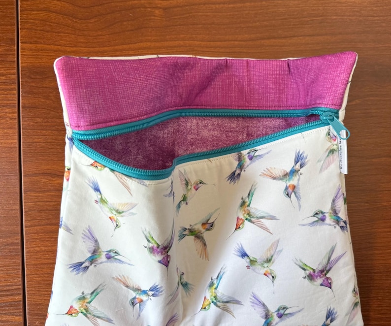 May include: A white fabric bag with a teal zipper and a magenta interior and trim. The bag is patterned with watercolor hummingbirds in flight, featuring shades of blue, green, purple, and orange. The bag is open, revealing the interior.