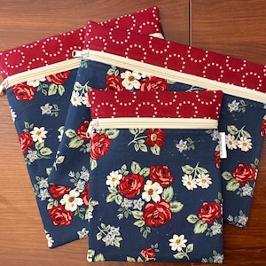 K&ouml;nnte beinhalten: Vier Rei&szlig;verschlusstaschen mit floralem Muster. Die Taschen haben einen marineblauen Hintergrund mit roten Rosen und wei&szlig;en Blumen. Der obere Teil und der Rei&szlig;verschluss sind cremefarben, und der obere Rand ist roter Stoff mit wei&szlig;en Sternakzenten.