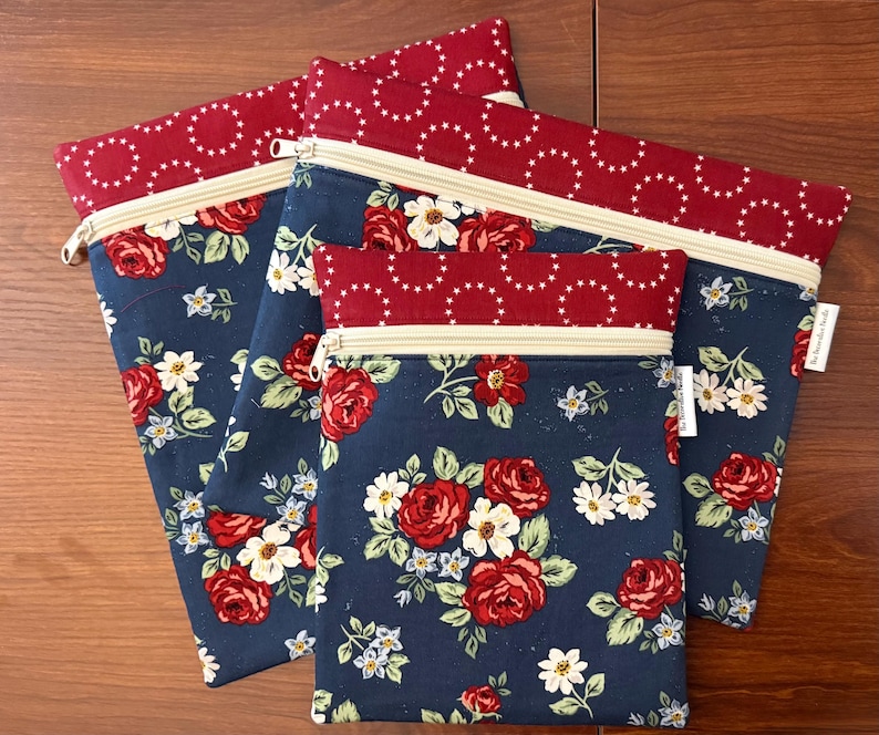 K&ouml;nnte beinhalten: Vier Rei&szlig;verschlusstaschen mit floralem Muster. Die Taschen haben einen marineblauen Hintergrund mit roten Rosen und wei&szlig;en Blumen. Der obere Teil und der Rei&szlig;verschluss sind cremefarben, und der obere Rand ist roter Stoff mit wei&szlig;en Sternakzenten.