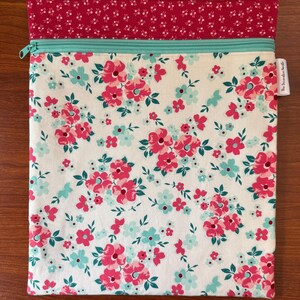 Puede incluir: Una bolsa rectangular de tela con estampado floral. La bolsa presenta un fondo blanco con flores rosas y turquesas. En la parte superior hay una tela estampada floral roja, con una cremallera turquesa. El texto "The Periwinkle Bundle" est&aacute; en una peque&ntilde;a etiqueta.