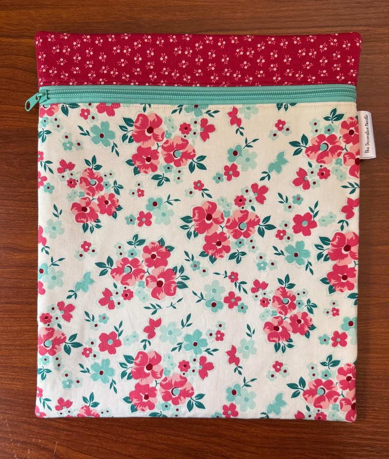 Puede incluir: Una bolsa rectangular de tela con estampado floral. La bolsa presenta un fondo blanco con flores rosas y turquesas. En la parte superior hay una tela estampada floral roja, con una cremallera turquesa. El texto "The Periwinkle Bundle" est&aacute; en una peque&ntilde;a etiqueta.