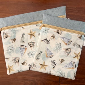 May include: Two zippered pouches with a nautical theme. The pouches feature a light blue top and a beige zipper. The main fabric is patterned with seashells, starfish, seahorses, and sailboats in shades of blue, beige, and brown.
