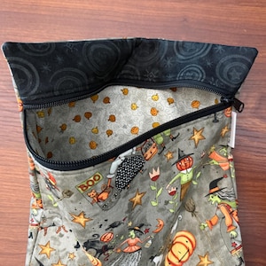 May include: A Halloween-themed zippered pouch with a gray exterior featuring orange pumpkins, witches, and stars. The interior lining is patterned with small orange pumpkins on a light gray background. The top edge is black with a swirl design.