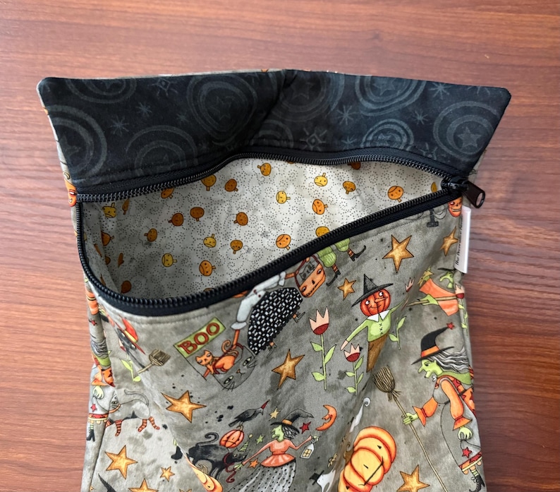 May include: A Halloween-themed zippered pouch with a gray exterior featuring orange pumpkins, witches, and stars. The interior lining is patterned with small orange pumpkins on a light gray background. The top edge is black with a swirl design.