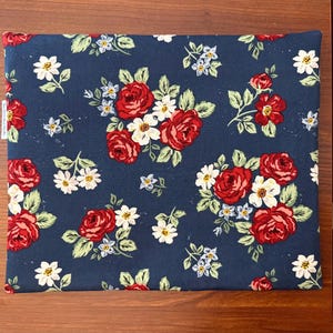 K&ouml;nnte beinhalten: Ein rechteckiges Stoffst&uuml;ck mit marineblauem Hintergrund, verziert mit einem Blumenmuster. Das Design zeigt rote Rosen, wei&szlig;e G&auml;nsebl&uuml;mchen und kleine blaue Blumen mit gr&uuml;nen Bl&auml;ttern. Der Artikel ist wahrscheinlich ein Kissenbezug oder ein &auml;hnliches Textil.