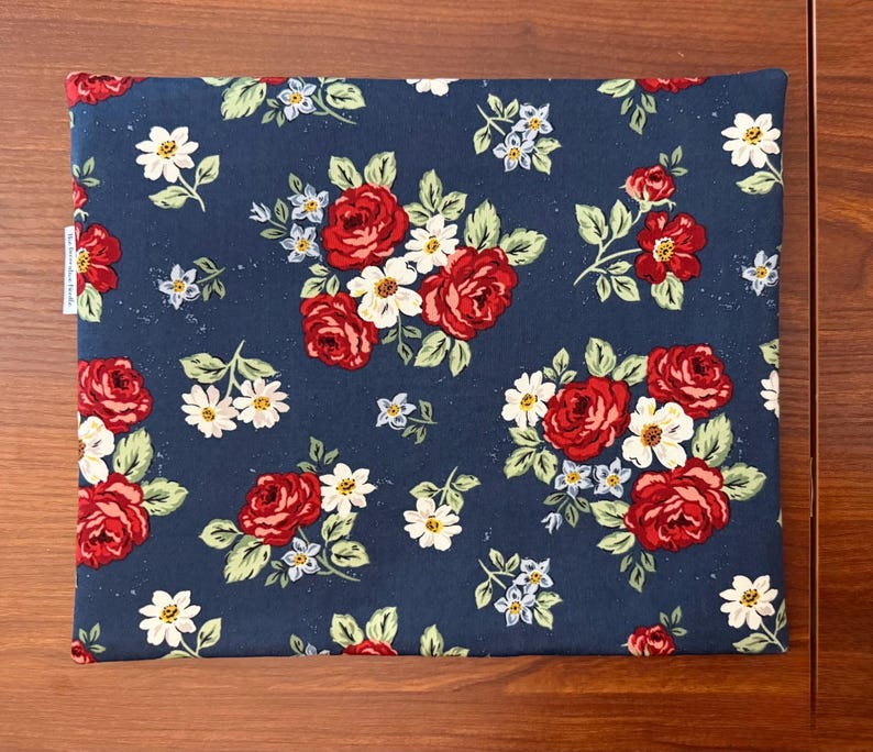 K&ouml;nnte beinhalten: Ein rechteckiges Stoffst&uuml;ck mit marineblauem Hintergrund, verziert mit einem Blumenmuster. Das Design zeigt rote Rosen, wei&szlig;e G&auml;nsebl&uuml;mchen und kleine blaue Blumen mit gr&uuml;nen Bl&auml;ttern. Der Artikel ist wahrscheinlich ein Kissenbezug oder ein &auml;hnliches Textil.