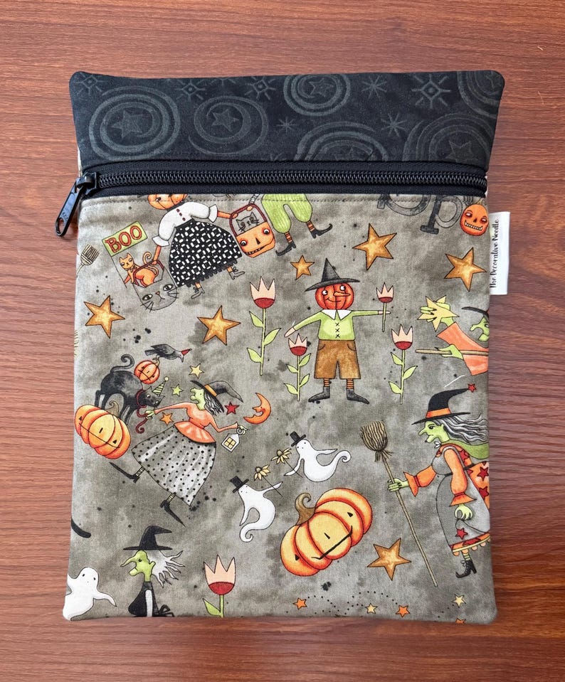 May include: A gray zippered pouch with a Halloween-themed pattern. The design features pumpkins, witches, ghosts, stars, and the word "BOO". The top section is black with a star and swirl pattern.