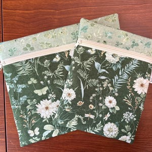 Pu&ograve; includere: Due pochette con cerniera con un motivo floreale verde scuro con fiori bianchi, farfalle e fogliame. La sezione superiore &egrave; verde chiaro con un design botanico simile. Le cerniere sono beige.