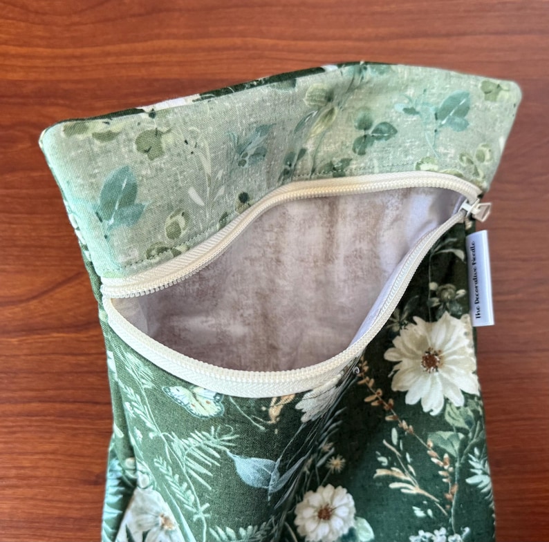 Pu&ograve; includere: Una pochette con cerniera floreale verde con una cerniera bianca. Il tessuto presenta un motivo di fiori bianchi, foglie e altra vegetazione su uno sfondo verde scuro. L'interno &egrave; di colore beige chiaro.