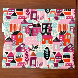 Puede incluir: Una bolsa de tela rectangular con un patr&oacute;n repetitivo de casas y &aacute;rboles estilizados en tonos vibrantes de rosa, naranja, rojo, negro y azul claro sobre un fondo crema. Una peque&ntilde;a etiqueta de tela blanca es visible en el borde izquierdo.
