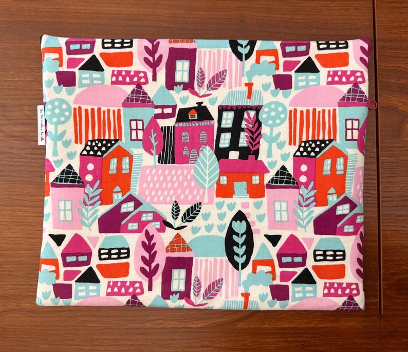 Puede incluir: Una bolsa de tela rectangular con un patr&oacute;n repetitivo de casas y &aacute;rboles estilizados en tonos vibrantes de rosa, naranja, rojo, negro y azul claro sobre un fondo crema. Una peque&ntilde;a etiqueta de tela blanca es visible en el borde izquierdo.