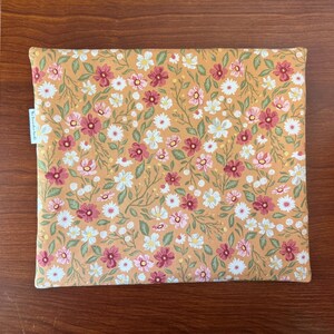 May include: A rectangular, floral-patterned fabric item, possibly a case or cover. The design features a mustard-yellow background with a repeating pattern of white, pink, and burgundy flowers with green leaves. A small white tag with text is visible on the left side.
