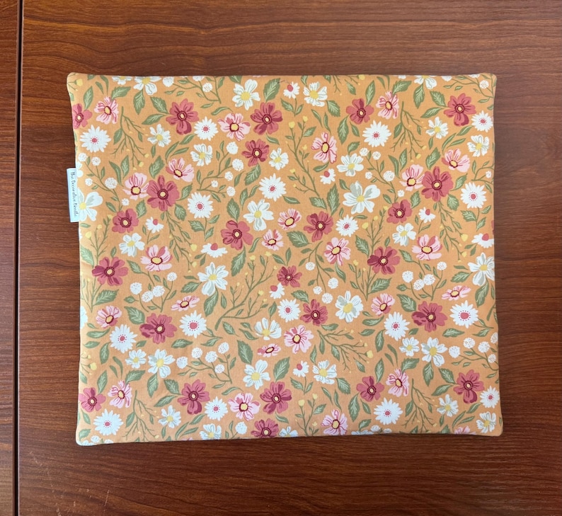 May include: A rectangular, floral-patterned fabric item, possibly a case or cover. The design features a mustard-yellow background with a repeating pattern of white, pink, and burgundy flowers with green leaves. A small white tag with text is visible on the left side.