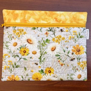 K&ouml;nnte beinhalten: Eine rechteckige Rei&szlig;verschlusstasche mit einem gelben Stoffband am oberen Rand und einem Hauptteil, das mit wei&szlig;en G&auml;nsebl&uuml;mchen, gelben Wildblumen und kleinen Bienen auf hellgrauem Grund mit feiner Schreibschrift bedruckt ist.