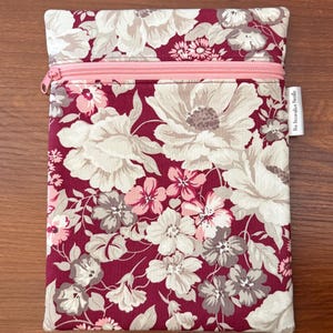 May include: A rectangular fabric pouch featuring a deep burgundy background with a floral pattern in shades of cream, light pink, and muted gray. The pouch includes a light pink horizontal zipper closure and a small white tag labeled The Decorative Needle.