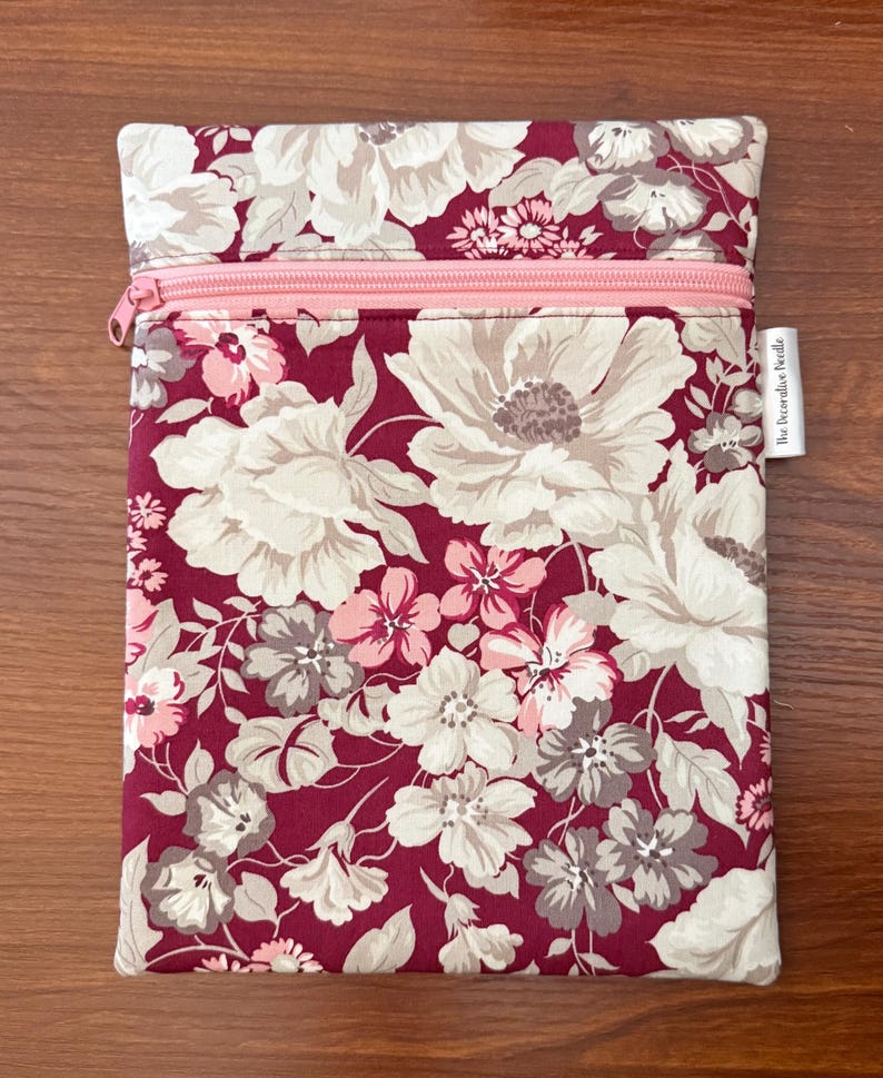 May include: A rectangular fabric pouch featuring a deep burgundy background with a floral pattern in shades of cream, light pink, and muted gray. The pouch includes a light pink horizontal zipper closure and a small white tag labeled The Decorative Needle.