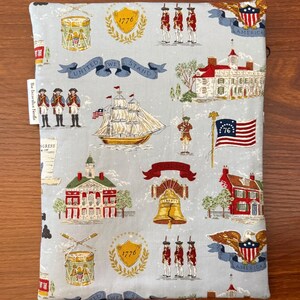 Op de afbeelding: Een lichtblauwe stoffen boekomslag met een patriottische print. Het ontwerp bevat illustraties van een schip, de Liberty Bell, een vlag en soldaten. De woorden "United We Stand" staan op een banner.