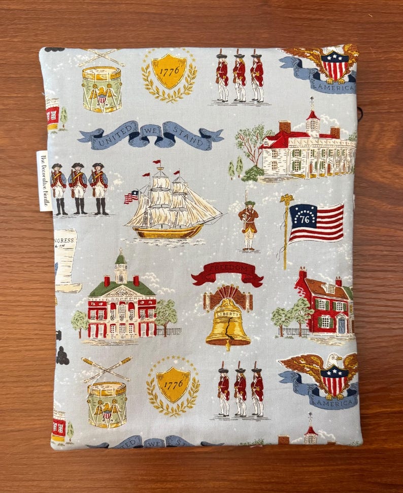 Op de afbeelding: Een lichtblauwe stoffen boekomslag met een patriottische print. Het ontwerp bevat illustraties van een schip, de Liberty Bell, een vlag en soldaten. De woorden "United We Stand" staan op een banner.