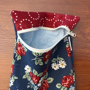 K&ouml;nnte beinhalten: Ein marineblaues T&auml;schchen mit einem Blumenmuster aus roten Rosen und wei&szlig;en G&auml;nsebl&uuml;mchen. Die obere Klappe ist tiefrot mit einem wei&szlig;en Sternmuster. Der Innenraum ist hellblau. Der Rei&szlig;verschluss ist wei&szlig;.