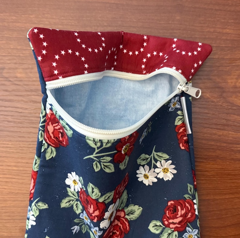 K&ouml;nnte beinhalten: Ein marineblaues T&auml;schchen mit einem Blumenmuster aus roten Rosen und wei&szlig;en G&auml;nsebl&uuml;mchen. Die obere Klappe ist tiefrot mit einem wei&szlig;en Sternmuster. Der Innenraum ist hellblau. Der Rei&szlig;verschluss ist wei&szlig;.