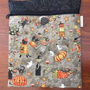 May include: A gray Halloween-themed zippered pouch with a black top and a black zipper. The pouch is covered in a pattern of witches, cats, pumpkins, stars, and ghosts. The word "BOO" is visible on the pouch.