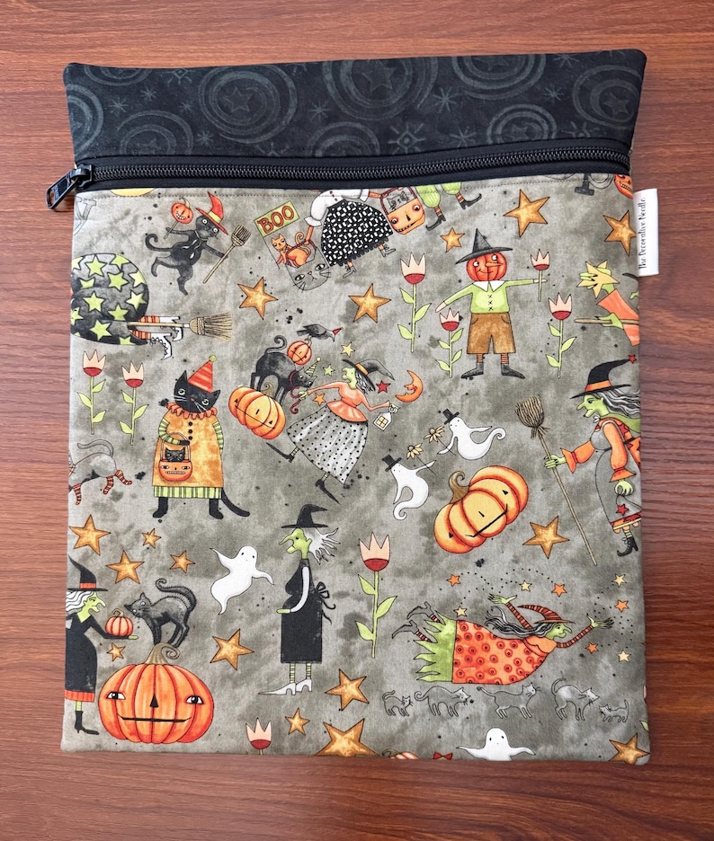 May include: A gray Halloween-themed zippered pouch with a black top and a black zipper. The pouch is covered in a pattern of witches, cats, pumpkins, stars, and ghosts. The word "BOO" is visible on the pouch.