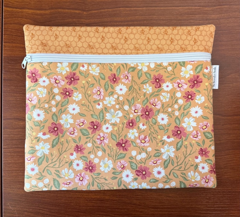 May include: A rectangular, floral-patterned pouch with a light blue zipper. The main body features a repeating pattern of pink, white, and burgundy flowers on a yellow background. The top section has a honeycomb pattern. The tag reads "The Decorative Beads".