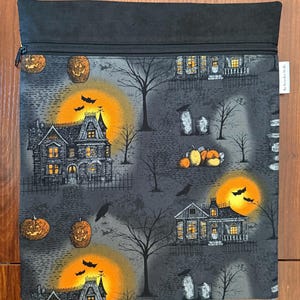 May include: A rectangular fabric pouch with a black zipper closure at the top. The pouch features a gray background with a Halloween-themed print of haunted houses, glowing orange full moons, jack-o'-lanterns, bare trees, and black bats.
