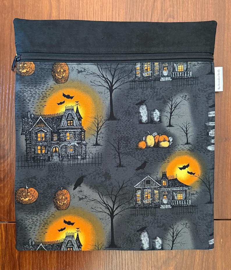 May include: A rectangular fabric pouch with a black zipper closure at the top. The pouch features a gray background with a Halloween-themed print of haunted houses, glowing orange full moons, jack-o'-lanterns, bare trees, and black bats.