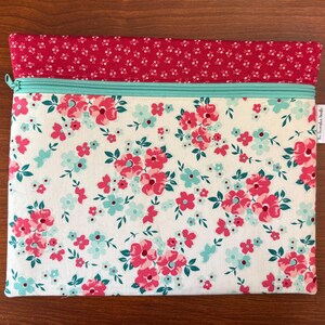 Puede incluir: Una bolsa con cremallera con estampado floral y una secci&oacute;n superior roja. La bolsa presenta un fondo blanco con flores rosas y azules y hojas verdes. La cremallera es de color verde azulado claro.