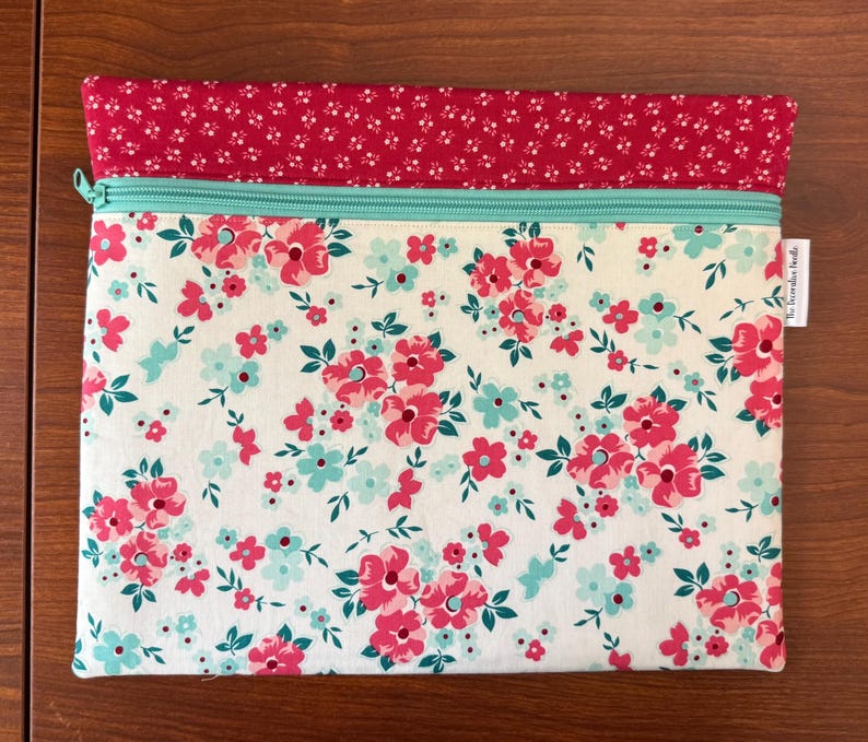 Puede incluir: Una bolsa con cremallera con estampado floral y una secci&oacute;n superior roja. La bolsa presenta un fondo blanco con flores rosas y azules y hojas verdes. La cremallera es de color verde azulado claro.