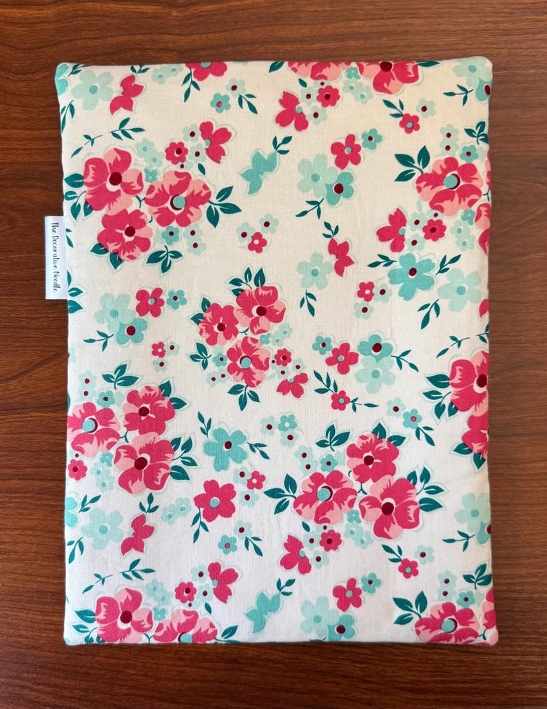 Puede incluir: Un art&iacute;culo de tela rectangular con un estampado floral. El dise&ntilde;o presenta un fondo blanco con flores rosas y turquesas y hojas verdes. El art&iacute;culo tiene una etiqueta que dice "The Decorative Needle."