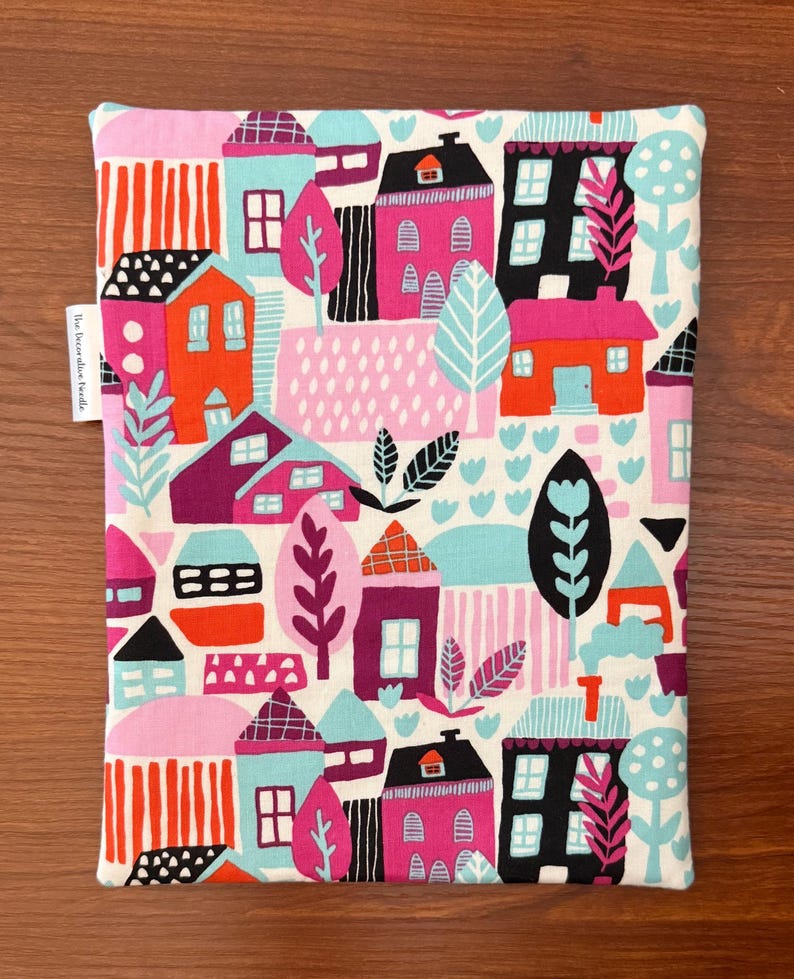 Puede incluir: Una bolsa de tela rectangular con un patr&oacute;n colorido de casas, &aacute;rboles y follaje en tonos rosa, naranja, negro y azul claro sobre un fondo blanco. Una peque&ntilde;a etiqueta de tela en el lateral muestra el texto The Decorative Needle.